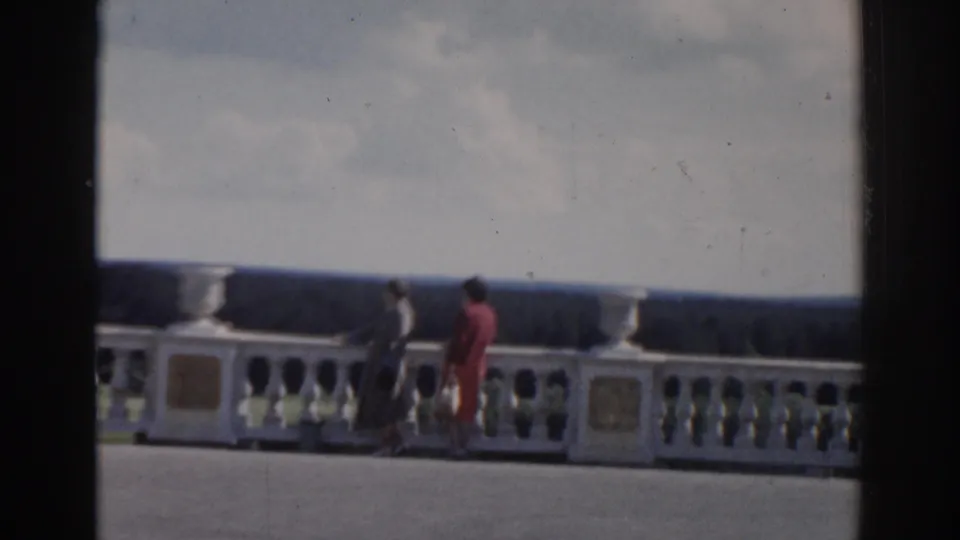 Two women in 1968 Moscow footage pause or walk beside the ornate white railing, framed by cloudy sky and a distant treeline.