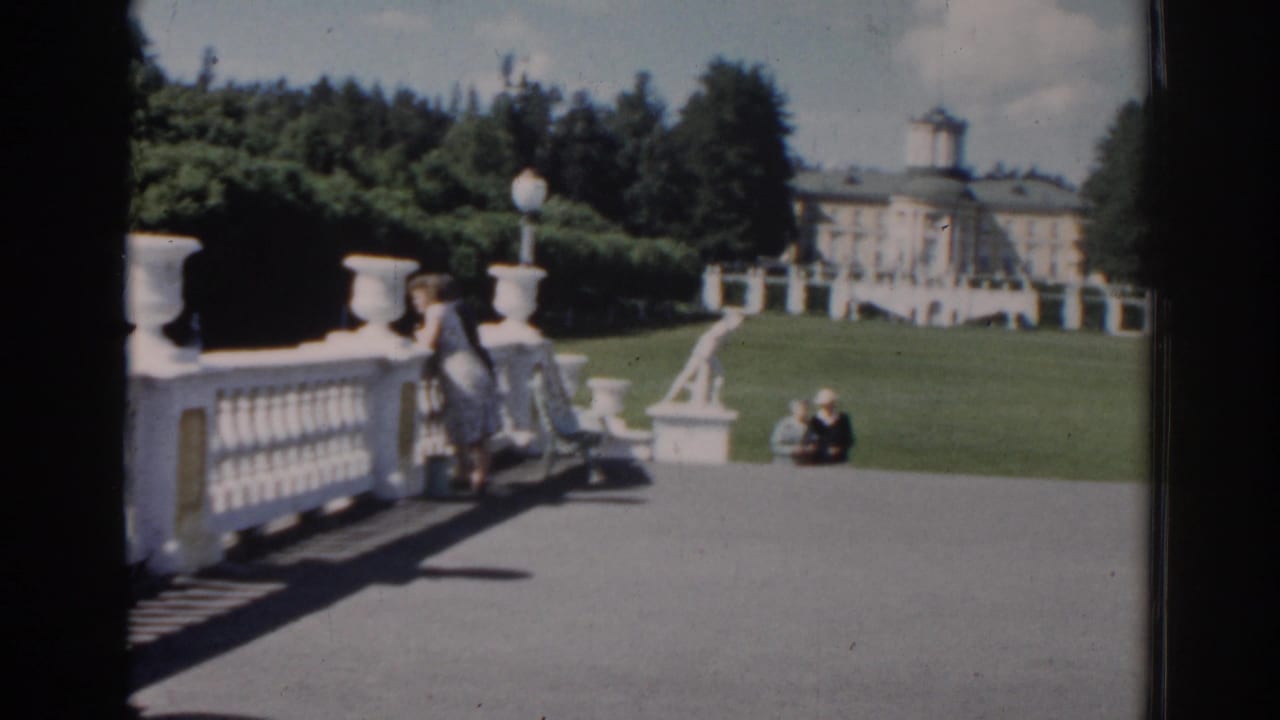 1968 Moscow home-movie frame showing a woman leaning near a white balustrade, with seated visitors, lawn, statues, and a palace-like building beyond.