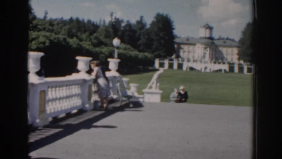 1968 Moscow home-movie frame showing a woman leaning near a white balustrade, with seated visitors, lawn, statues, and a palace-like building beyond.