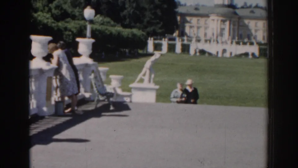 1968 Moscow park scene with one woman at the balustrade and another in a hat near the lawn, backed by statues and a classical palace-like facade.