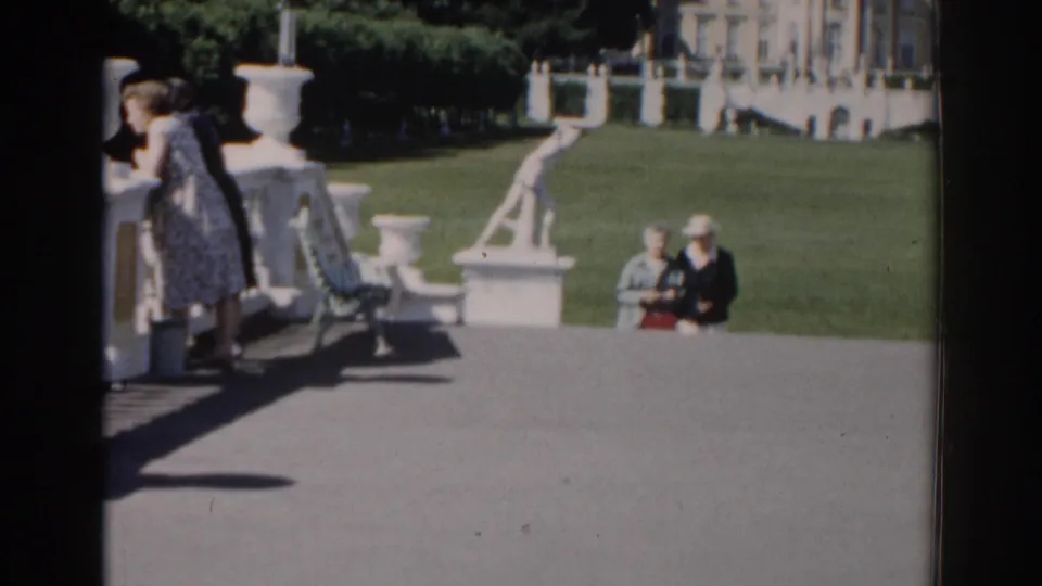1968 Moscow home-movie frame with a woman partly at the terrace edge, two visitors below, white garden sculpture, lawn, and classical architecture behind.