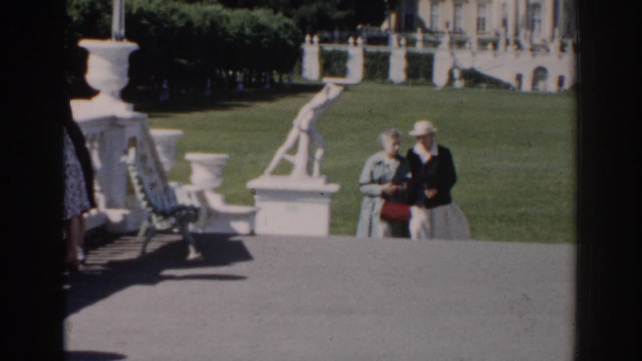 Elderly couple walking in a formal garden with a white statue and urns nearby.