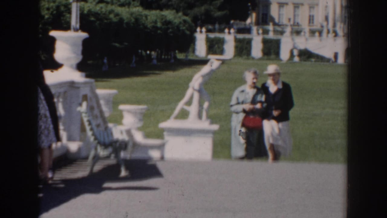 The couple continues toward the camera in a Moscow formal garden, the woman carrying a red handbag beside white statuary and clipped greenery.