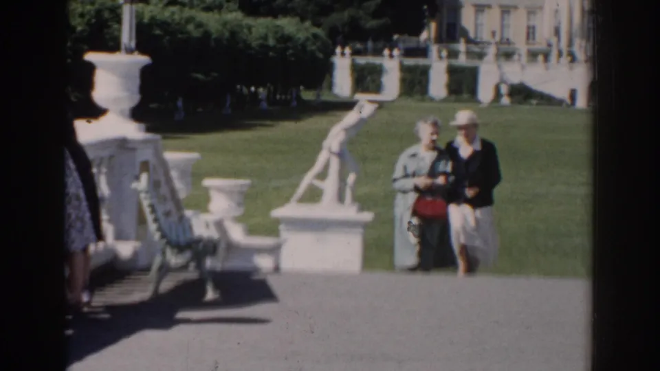 The couple continues toward the camera in a Moscow formal garden, the woman carrying a red handbag beside white statuary and clipped greenery.