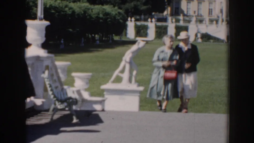 The woman in a light coat and her companion in a dark jacket walk near the camera in sunlit 1968 Moscow garden footage.