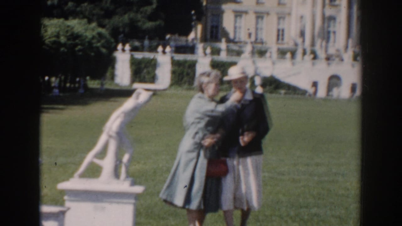 Two women stand on a lawn near a white sculpture and large building facade.