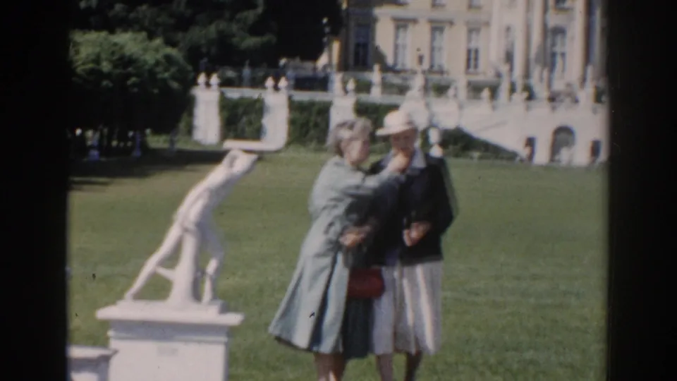 In 1968 Moscow home-movie footage, two women stand in a garden near a white classical sculpture and a large building facade.