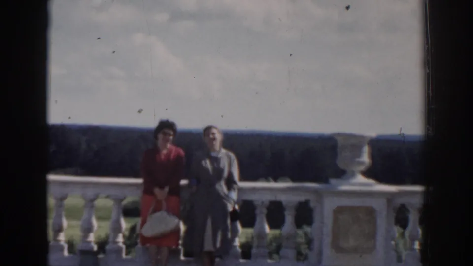 Two women in period dresses or coats stand against a stone balcony rail in Moscow, with a forested view beyond.