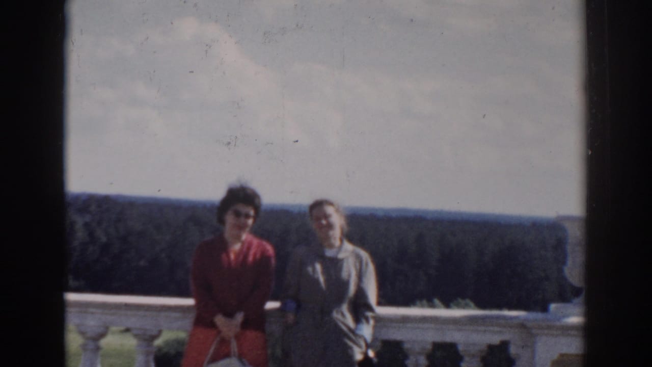 Two women seated or leaning by a stone balcony with forest and sky behind them.