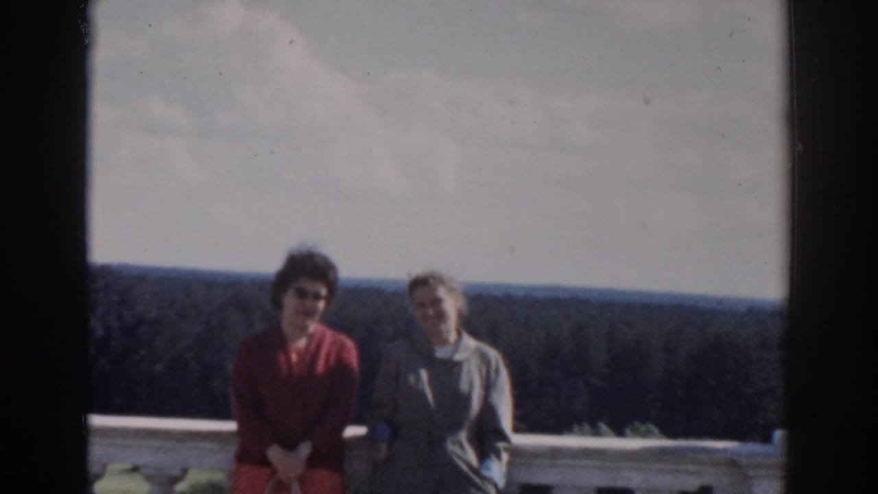 Two women remain framed at a stone balcony overlook, one in red and one in a light coat, against a wooded Moscow landscape.