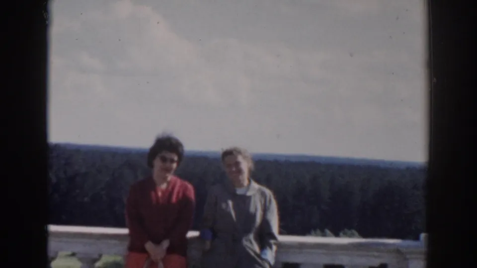 Two women remain framed at a stone balcony overlook, one in red and one in a light coat, against a wooded Moscow landscape.