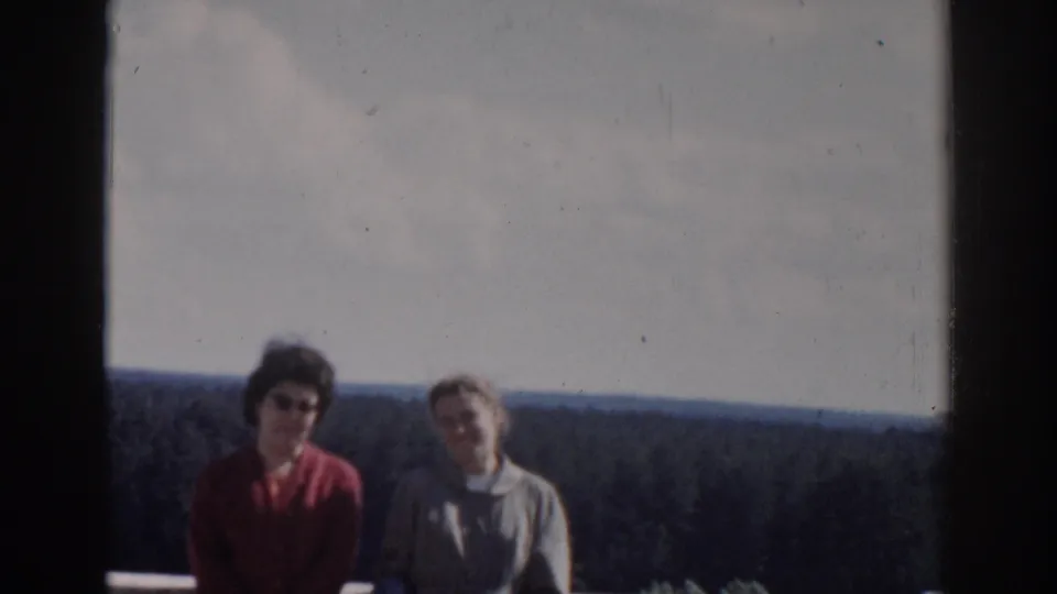 Handheld 8mm frame of two women on a balcony overlook, softly exposed with trees and open sky behind them.