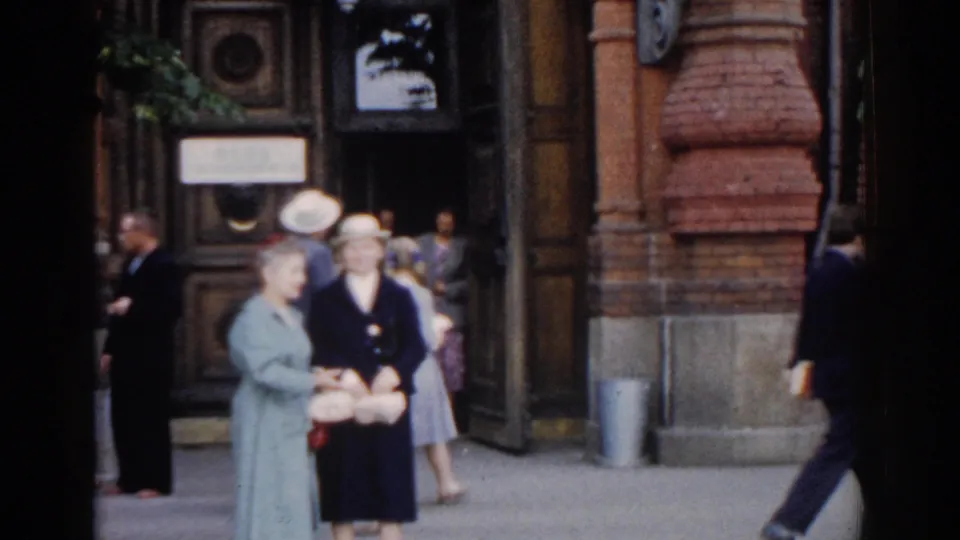 Street scene in Moscow home-movie footage showing two women near an entrance, with pedestrians and a red brick facade nearby.