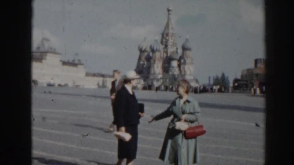 Two women in coats appear closer to the camera in Red Square, with St. Basil's Cathedral, pedestrians, and open paving visible.
