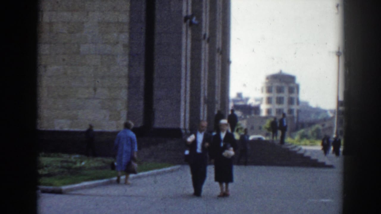 Pedestrians walk beside a tall masonry building with a distant domed structure visible.