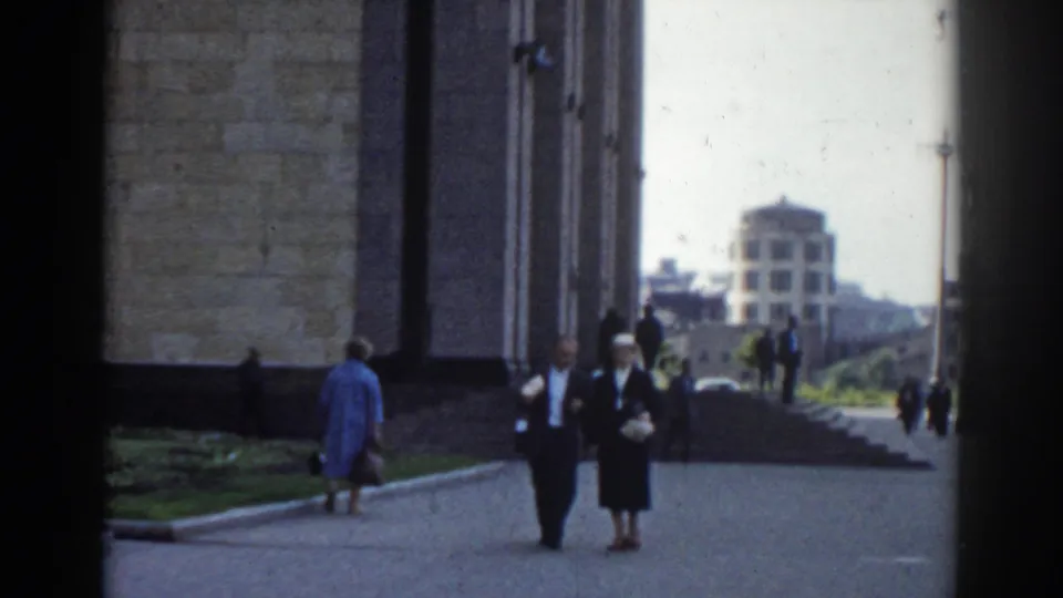 Pedestrians move along the edge of a large masonry building in Moscow, with a domed structure and open walkway in the distance.