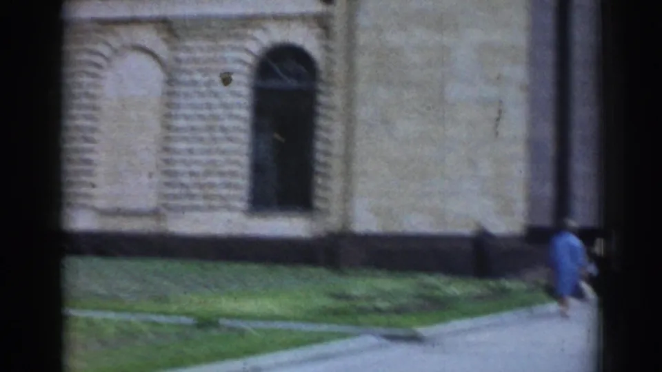 A handheld 8mm view lingers on the brick and stone wall, arched window, grass strip, and a pedestrian near the building.