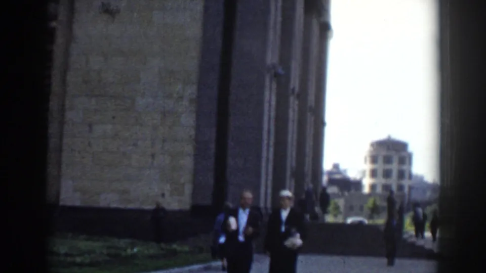 An older couple walks past the camera along a Moscow building facade, with other pedestrians and a domed building visible down the walkway.