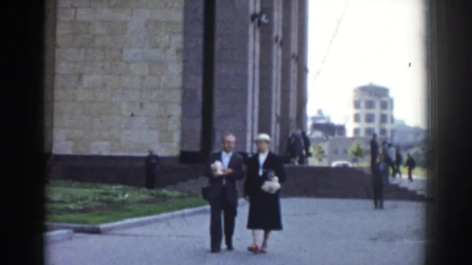 In metadata-supported 1968 Moscow, a man and woman walk across a paved courtyard beside a large stone building.