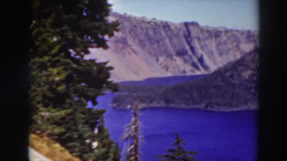 1937 8mm view of Crater Lake, Oregon, framed by dark evergreens with blue water and pale volcanic cliffs in the distance.