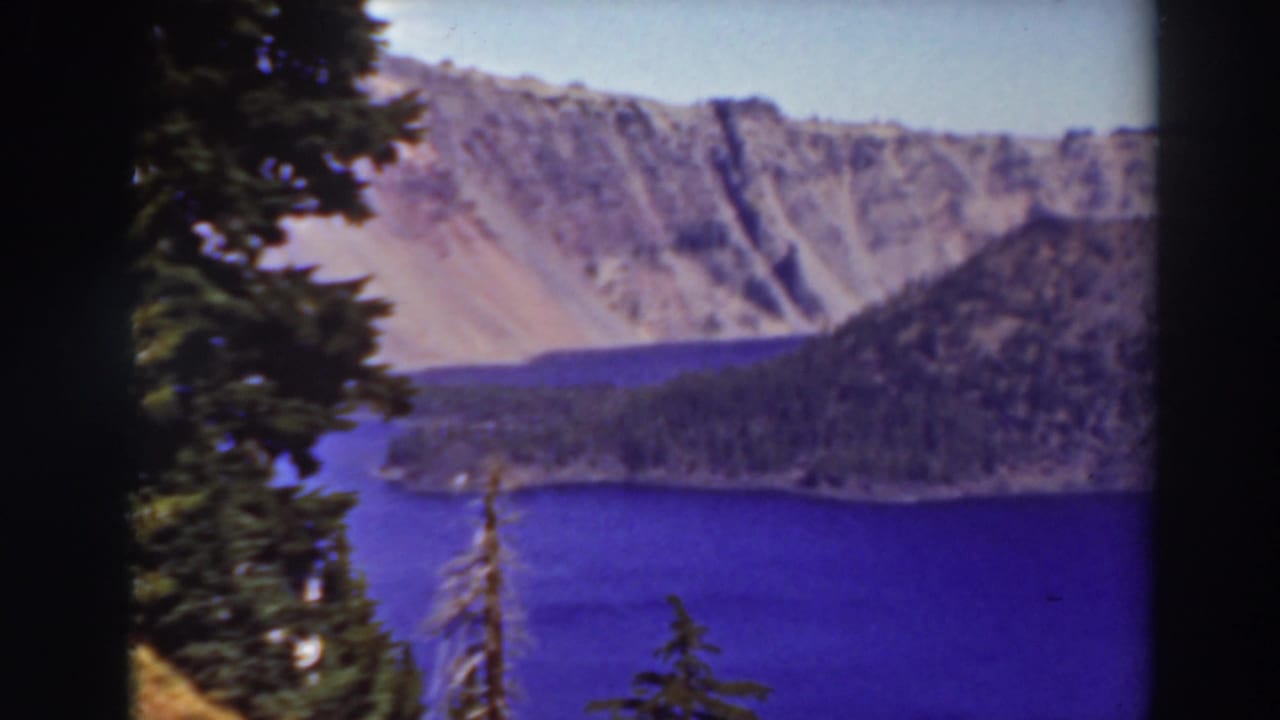 Blue lake, evergreen trees, wooded slope, and rocky crater rim under a clear sky.