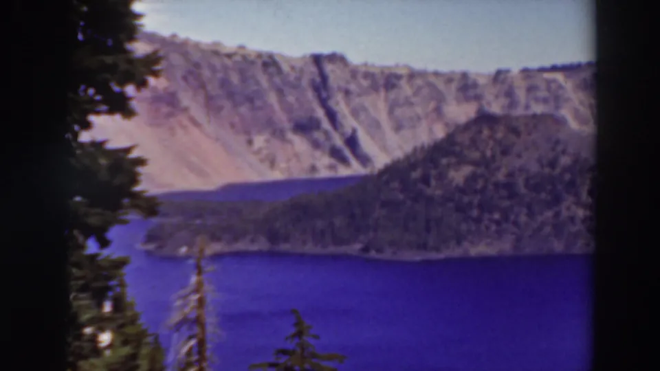 1937 8mm frame of Crater Lake, Oregon, with foreground pines, saturated blue water, a dark wooded ridge, and pale cliff walls.