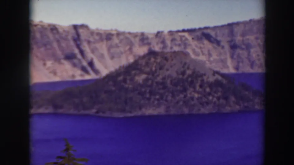 1937 Crater Lake home-movie frame showing Wizard Island centered against the caldera rim with saturated blue water in the foreground.