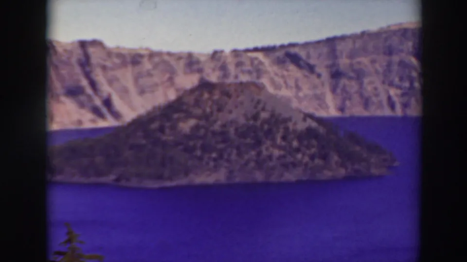 1937 8mm frame of Wizard Island in Crater Lake, with purple-blue water, pale caldera cliffs, and a dark evergreen at the lower edge.