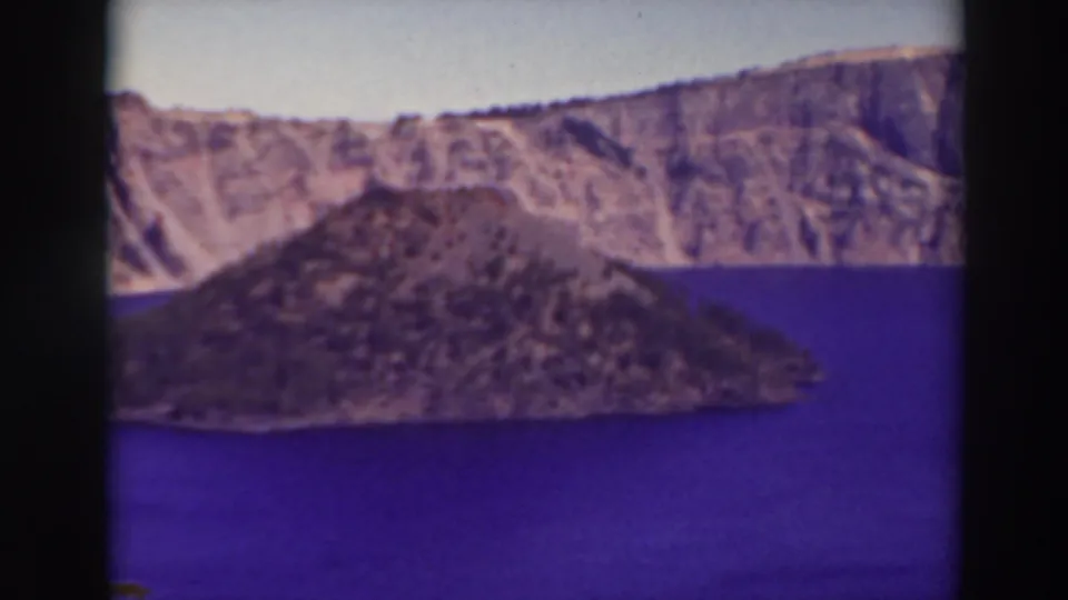 Late frame from a 1937 Crater Lake 8mm shot, showing Wizard Island, blue water, and the steep caldera wall under a pale sky.
