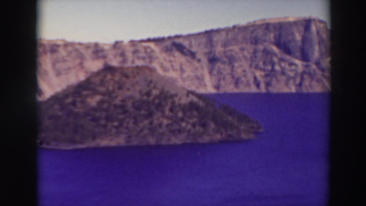 1937 Crater Lake aerial view with deep blue water, a rocky volcanic island, and rugged caldera wall framed by 8mm film borders.