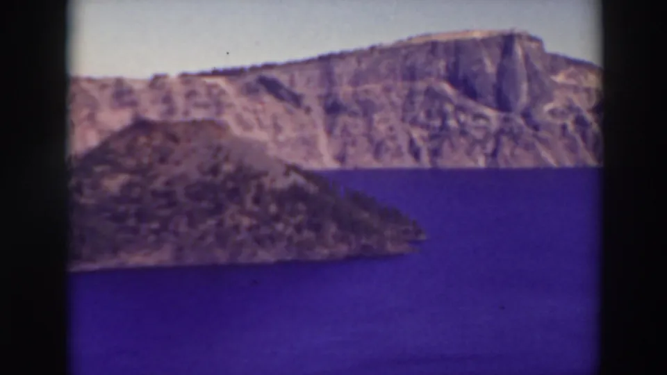 Aerial 8mm view at Crater Lake showing blue water, a rocky island form, and the steep caldera wall in soft color.