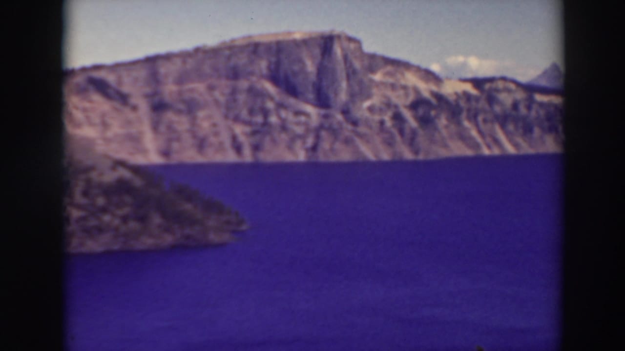 1937 8mm view of Crater Lake, Oregon, showing deep blue water, caldera cliffs, and a dark foreground slope.