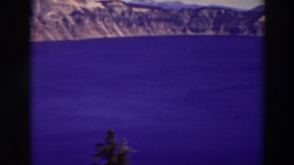 1937 8mm view of Crater Lake, Oregon, with saturated blue water, distant volcanic rim cliffs, and a small evergreen near the lower frame.