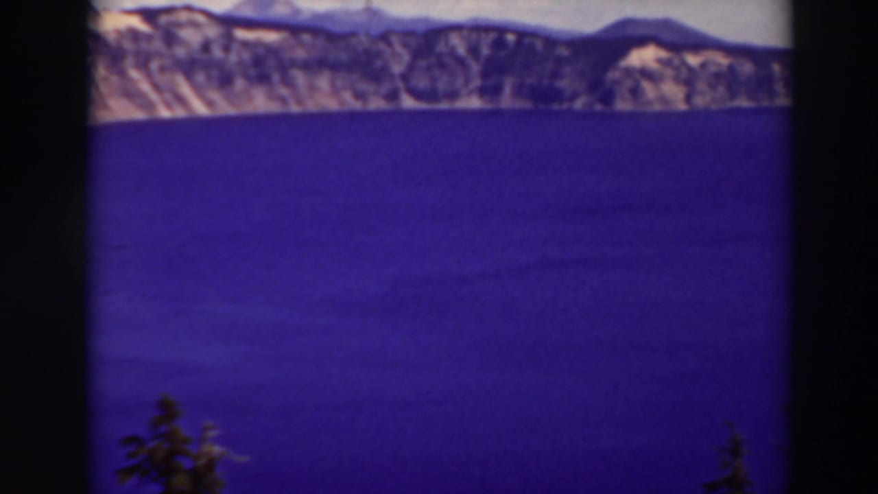 Blue lake surface below distant cliffs, with dark film borders and a small evergreen at bottom left.