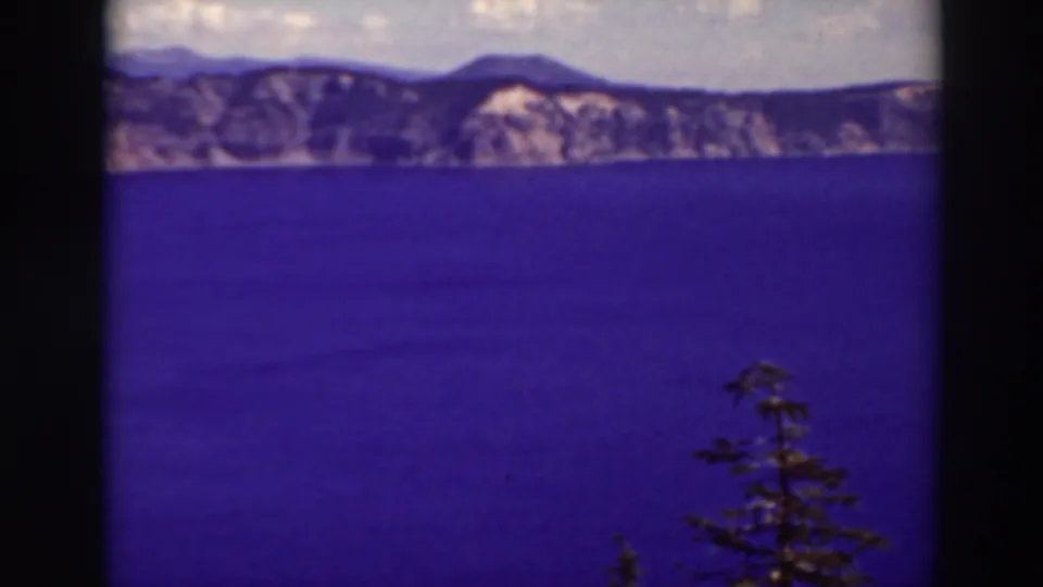 A 1937 8mm frame looks across Crater Lake, Oregon, toward volcanic rim cliffs, with blue water and evergreen branches in the foreground.