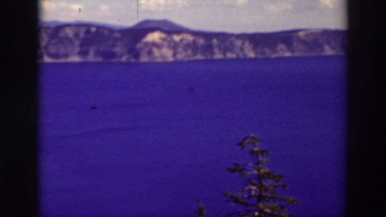 1937 8mm home-movie frame of Crater Lake, Oregon, showing blue water, distant cliffs, cloudy sky, and a foreground evergreen.