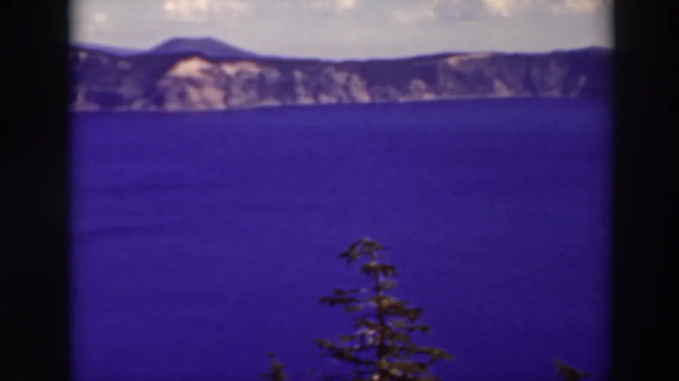 Crater Lake, Oregon, appears in a soft 1937 8mm frame with saturated blue water, rim cliffs, clouds, and a foreground tree.
