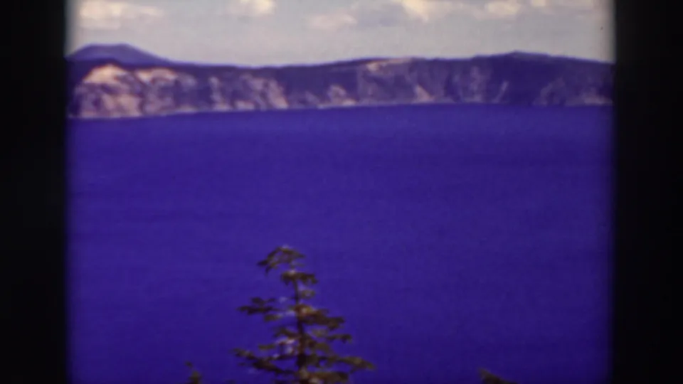 1937 Crater Lake home-movie frame showing blue-purple water, pale rim cliffs, cloudy sky, and a small evergreen foreground shape.