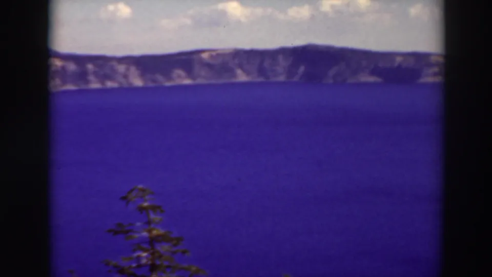 Soft 8mm frame from Crater Lake, Oregon, with deep blue water, dark volcanic rim, cloudy sky, and an evergreen at the bottom.