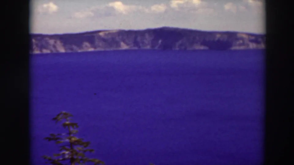 1937 8mm view of Crater Lake with blue water, rim cliffs, cloudy sky, and a small evergreen at the lower left edge.