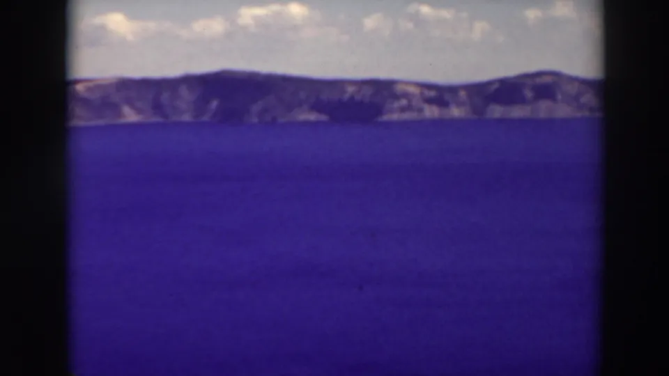 A 1937 8mm frame shows Crater Lake’s blue surface below the distant rim cliffs and a pale, clouded sky.