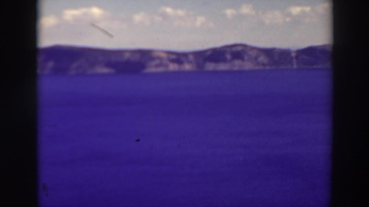 1937 8mm home-movie view of Crater Lake, Oregon, with blue water, distant rim slopes, pale clouds, and visible film gate edges.