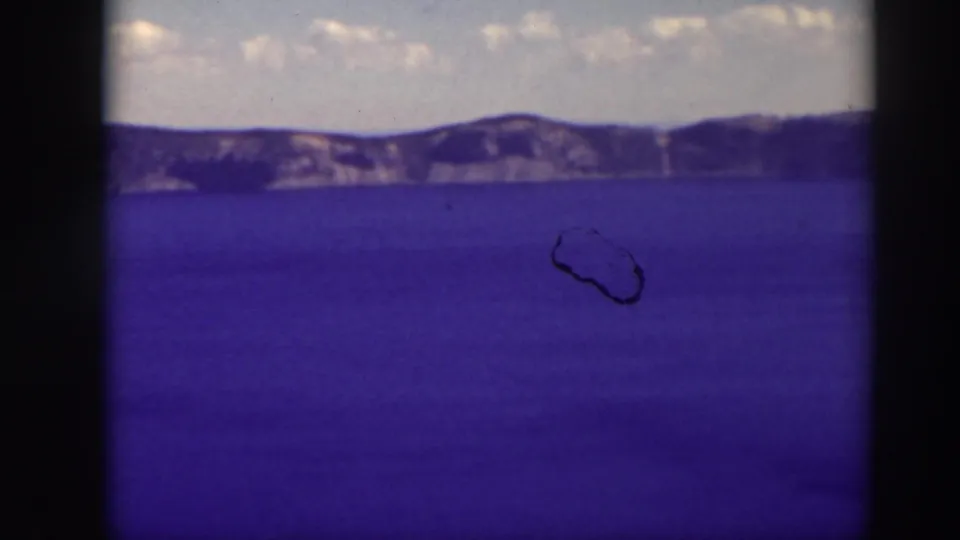 Crater Lake, Oregon, appears across a 1937 8mm frame, with saturated blue water, rim cliffs, clouds, and a visible dust loop.