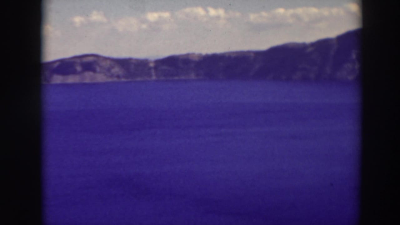 1937 8mm view of Crater Lake, Oregon, showing blue water and distant rim cliffs beneath scattered clouds.
