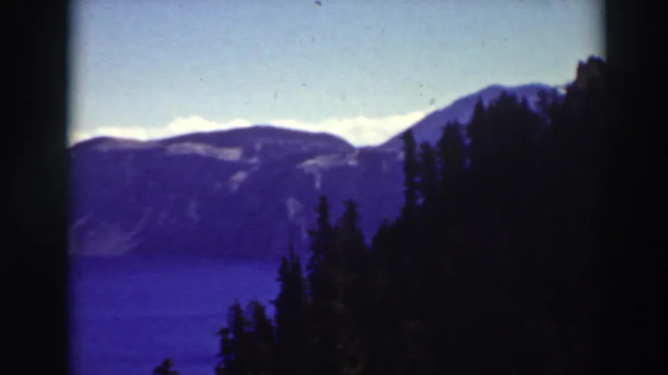 Crater Lake, Oregon, appears beyond dark evergreen silhouettes, with blue water and misty ridges in a soft 1930s 8mm frame.
