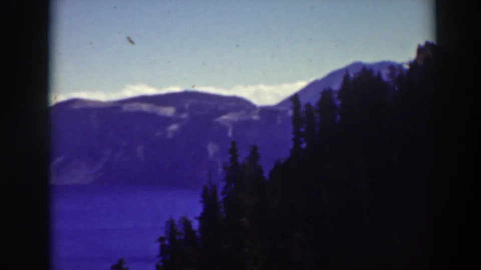 Soft 8mm frame of Crater Lake with blue water, hazy mountain ridges, pale sky, and dense dark evergreens along the right side.