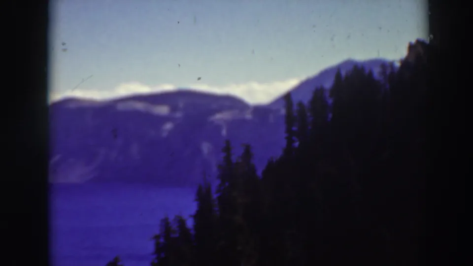 Crater Lake landscape in a 1937 home-movie frame, showing dark conifers, blue water, distant ridges, pale sky, and visible film wear.