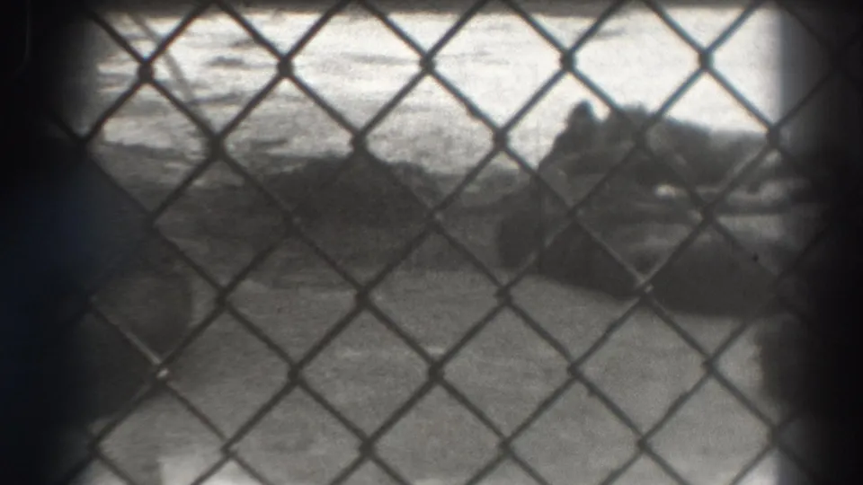 Black-and-white 8mm frame with chain-link fence foreground and indistinct sand or shoreline forms beyond it.