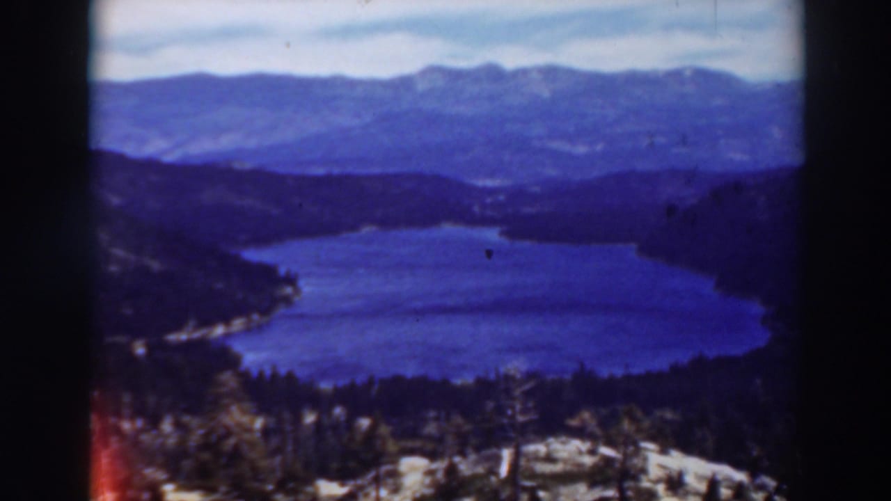 1937 Lake Tahoe home-movie frame showing blue water below dark wooded hills and distant mountain ranges under a pale sky.