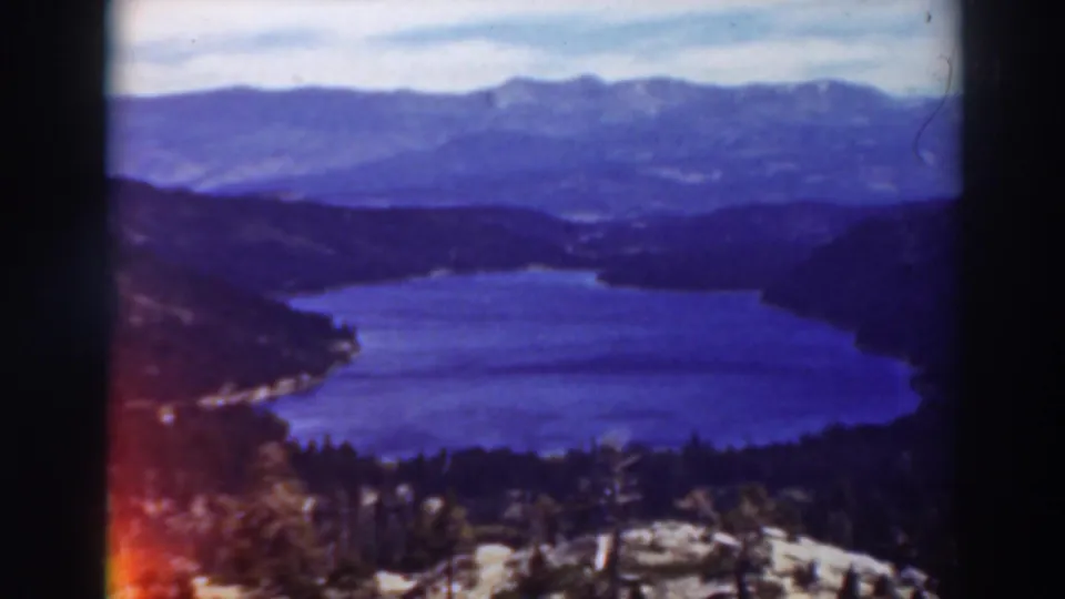 1937 Lake Tahoe frame with the lake centered between wooded slopes, distant mountains, cloudy sky, and 8mm edge coloration.